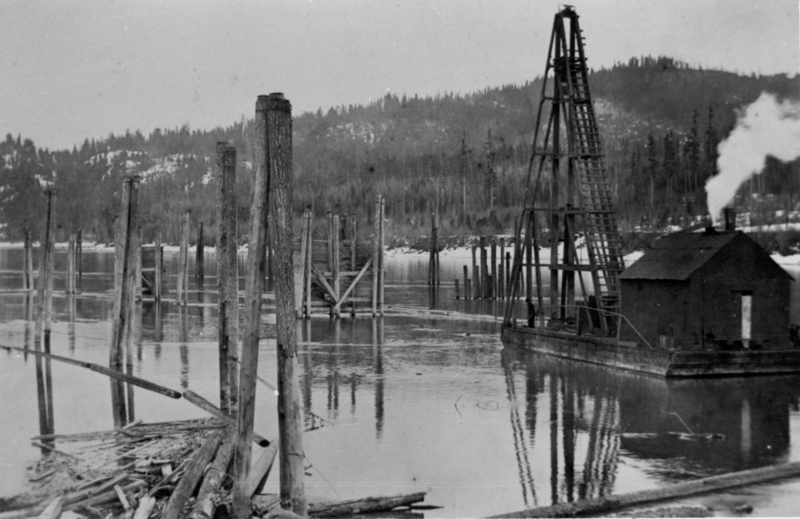A large river or lake with numerous wooden pilings sticking out of the water. To the right, there is a structure on a barge, emitting smoke, with a tall, crane-like framework beside it. In the background, there are forested hills.