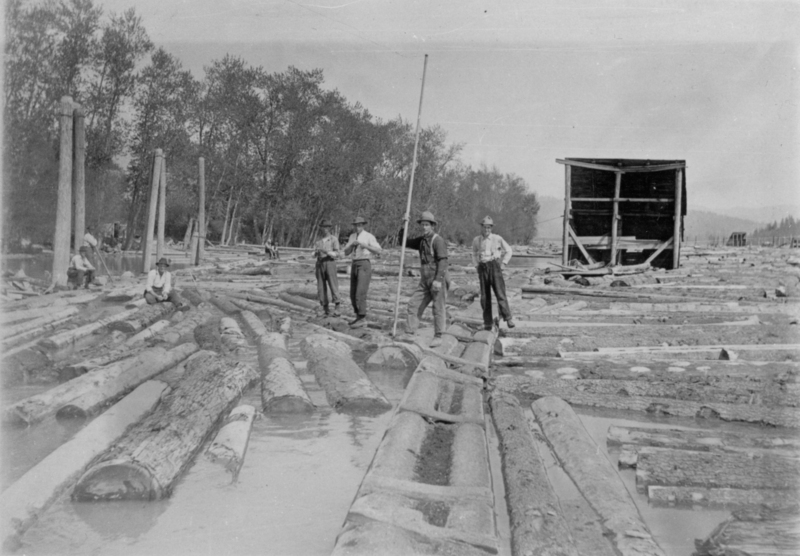 Several men stand on floating logs in a watery area, one holding a long pole. Trees and a large wooden structure are visible in the background. Logs cover the water surface extensively.