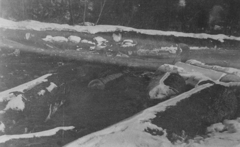 Logs partially covered in snow lying on the ground near a snowy embankment. A person is standing in the background, possibly holding a tool. Trees are visible in the distance.