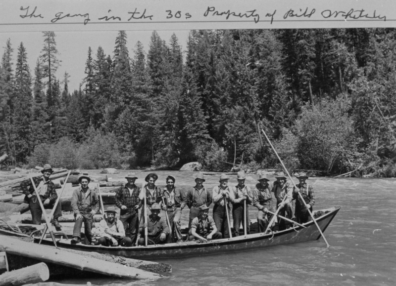 A group of men standing and sitting in a boat on a river, surrounded by logs and paddles. Behind them, dense trees line the riverbank. Handwritten text near the top reads: "The gang in the 30s Property of Bill..."