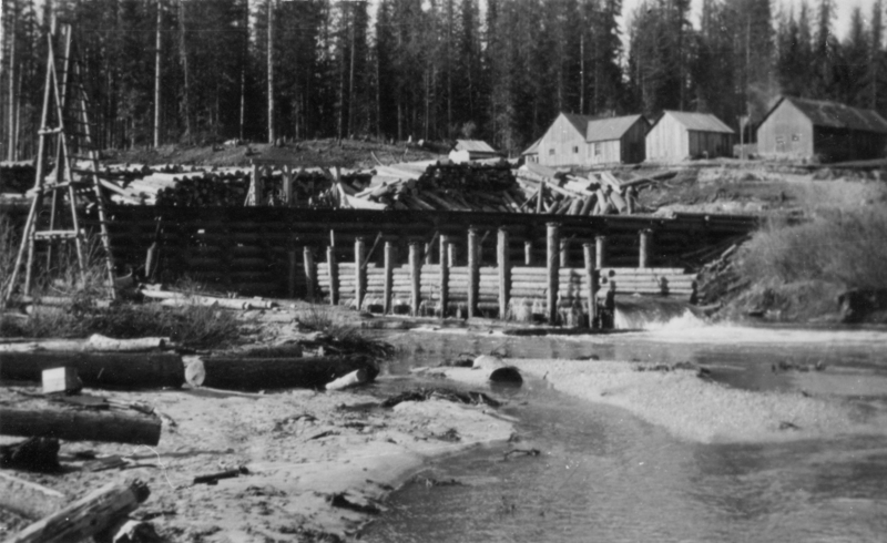 A wooden structure, possibly a dam, spans a stream, surrounded by logs. In the background, there are several small wooden buildings and tall trees.