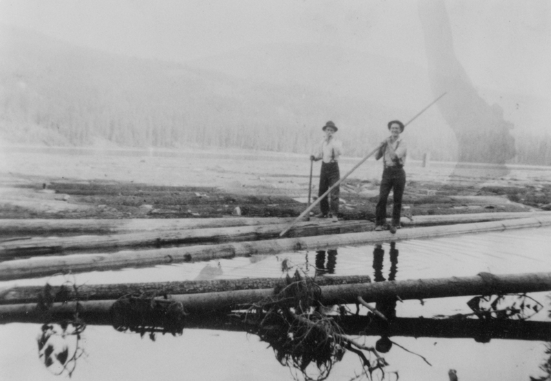 Two men standing on logs floating on a body of water, both holding long poles, with a backdrop of trees in the distance.