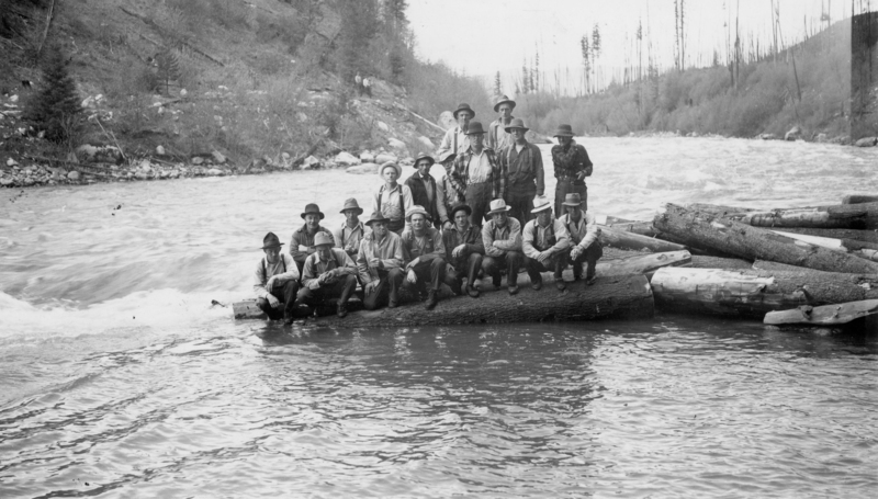 A group of people wearing hats and outdoor clothing stand and sit on logs near a flowing river. There is a wooded hillside in the background.