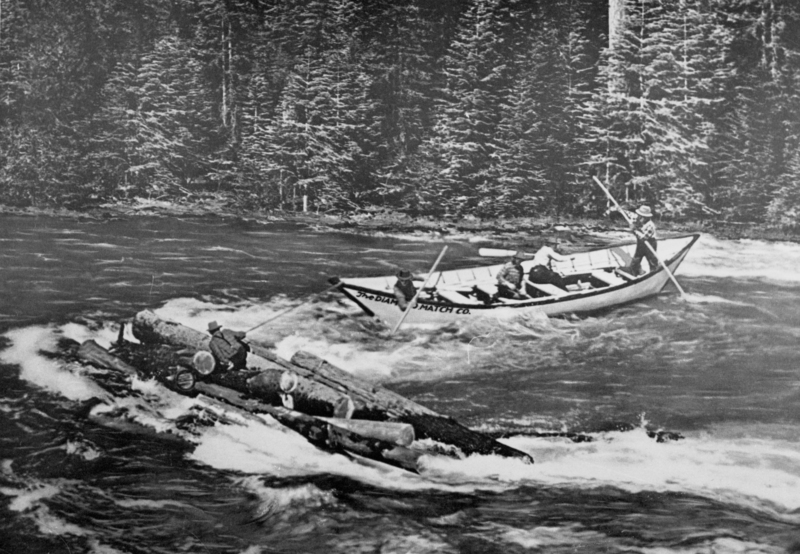 A man crouching on a raft of logs in a river, with another boat nearby rowing through the water. Trees line the riverbank in the background. The text visible on the side of the boat reads: "THE DIAMOND MATCH CO."