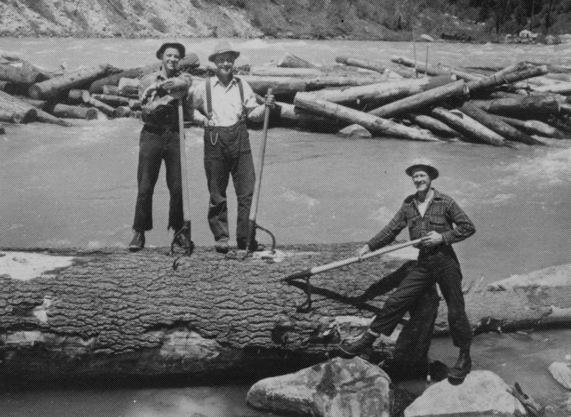 Three men standing on large logs in a river, each holding a long tool. Logs and rocks are visible in the background.