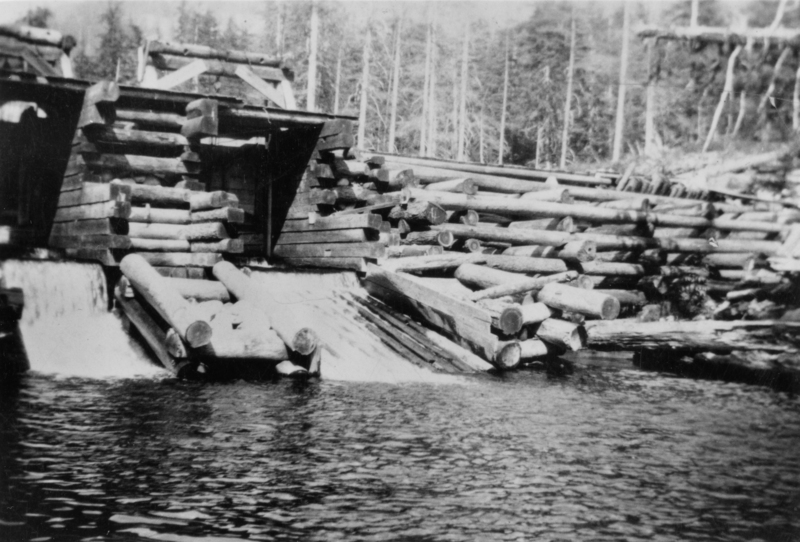 A wooden dam stretching across a body of water, with water flowing beneath it. Logs are stacked on top of the structure.