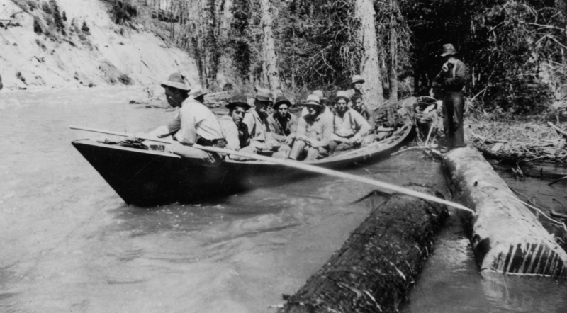 A group of people sitting in a canoe on a river, with one person standing on a log nearby. The surrounding area is wooded and the water is flowing gently.