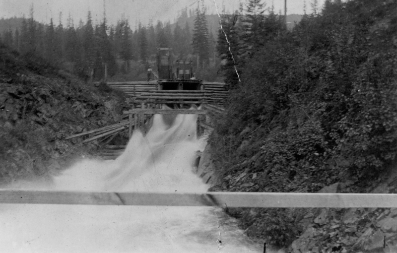 A wooden structure with rushing water flowing beneath it, surrounded by rocky terrain and dense trees in the background. A person stands on the structure above the water.