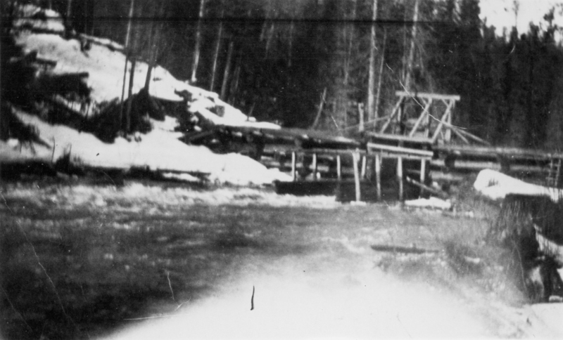 A river flowing in the foreground with a wooden bridge structure spanning across it. Snow is visible on the banks, and tall trees stand in the background.
