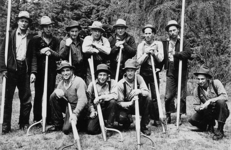A group of eleven men standing and kneeling outdoors, each holding a long-handled tool. They are wearing hats and work clothes, and there are trees in the background.