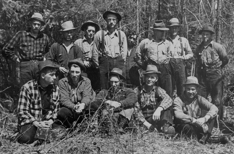 A group of men wearing hats and plaid shirts, standing and sitting in a wooded area. There are several trees and branches in the background. Some of the men are seated on the ground while others stand behind them.