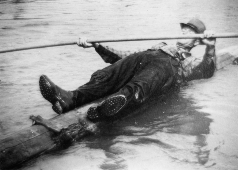 A person lying on a log in water, holding a long pole and wearing a hat. They are dressed in rugged clothing and boots with a spiked sole.