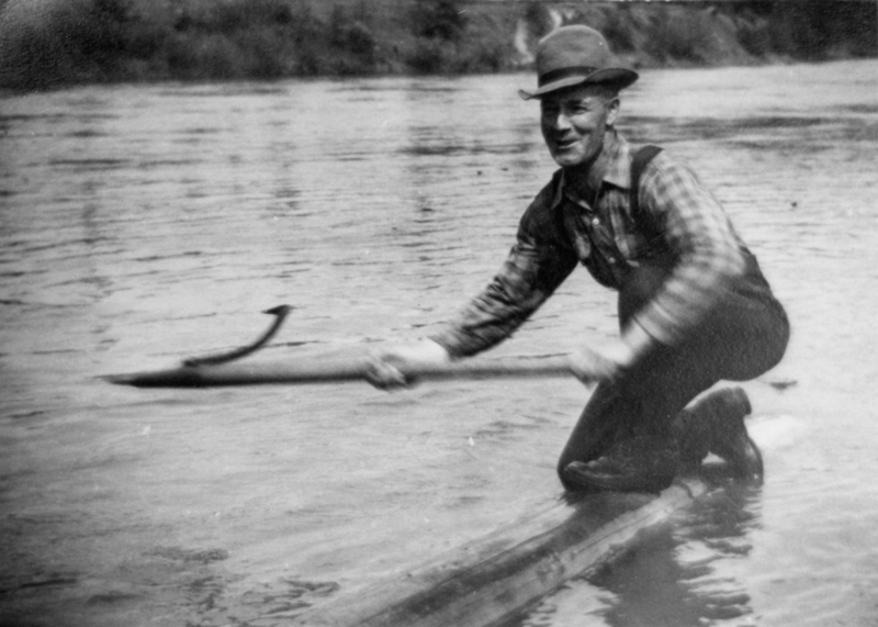 A man wearing a hat and a plaid shirt is kneeling on a log in a body of water, holding a long stick or pole.