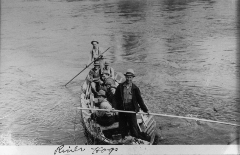 A group of men are in a boat on a river. Some are sitting while one man stands at the back holding a long oar. The boat is pointed upstream, with a few men wearing hats. The water is calm. Handwritten text at the bottom of the image reads: "River Hogs."