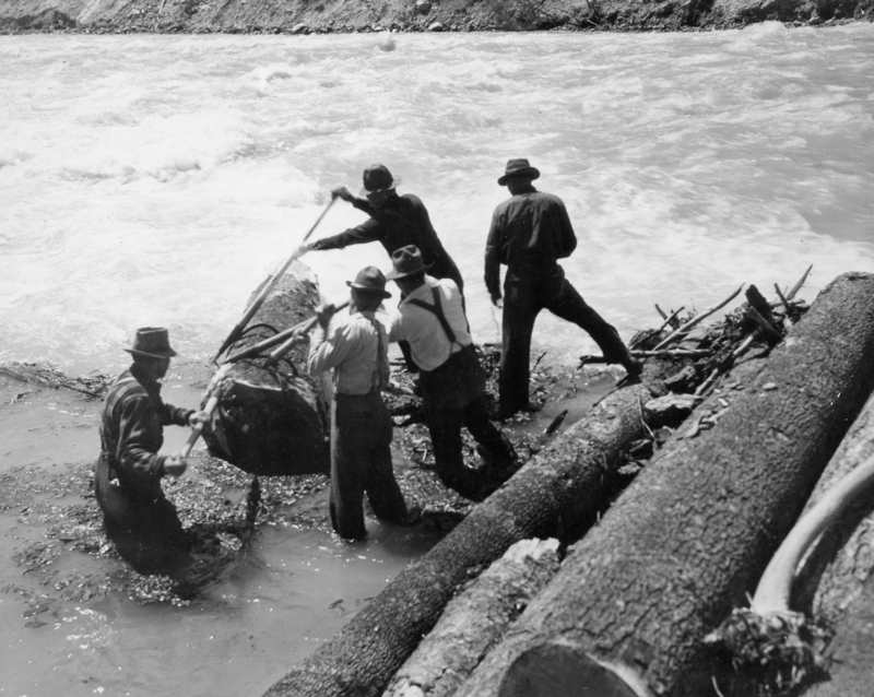 Five men wearing hats and work clothes are standing at the edge of a river. They are using poles to maneuver large logs in the water. Some logs are piled beside them on the riverbank.