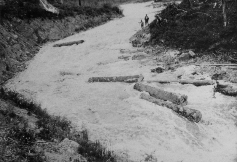 A river with logs floating in it, bordered by steep banks, with two people standing on the right side of the bank in the background.