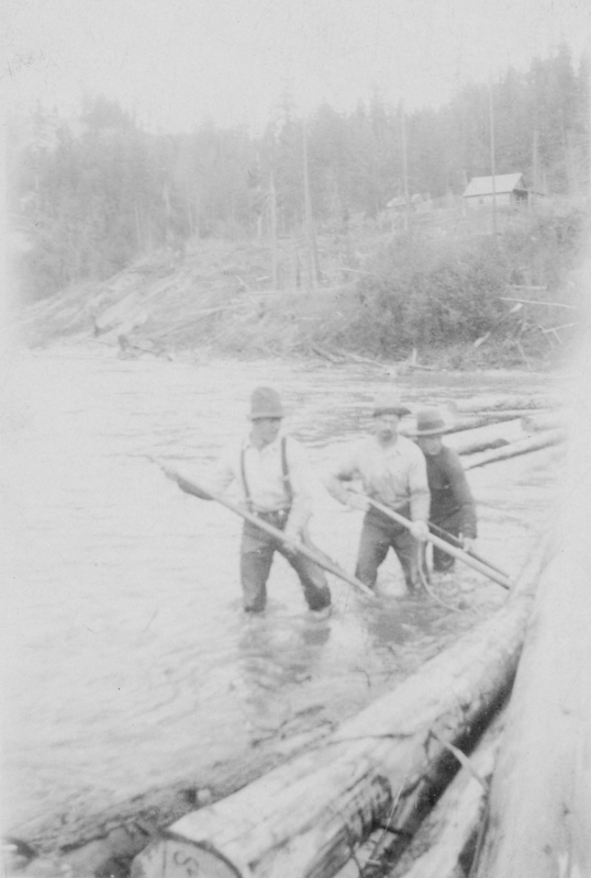 Three men standing in a river using poles to manage logs. Logs float in the water beside them. Trees and a wooden building are visible in the background.