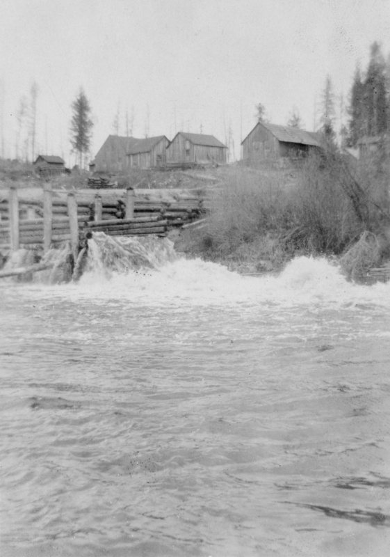 A turbulent stream flows beneath a wooden log dam with a collection of wooden buildings and sparse trees in the background.