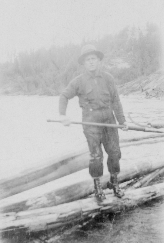 A person stands on logs by a river, holding a long pole. They are dressed in outdoor clothing, including a hat and boots. Trees are visible in the background.