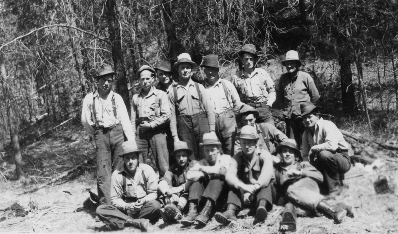 A group of men in work clothing and hats gathered together in a forested area, with some standing and others sitting or crouching on the ground. The background consists of trees and natural vegetation.