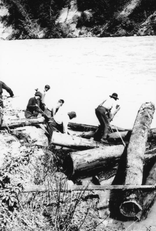 Several people are standing on large logs by a riverbank. They are wearing hats and work clothes, and the landscape includes water and tree-covered hills in the background.