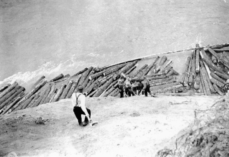 A man wearing a hat and suspenders stands on a sloped embankment near the water's edge. Below, several logs are floating, and two horses are visible next to the logs.