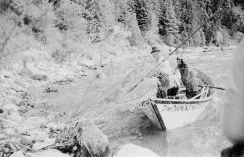 A small group of people is seated in a boat navigating a river. The boat is close to the rocky riverbank, and there are trees and shrubs in the background. One person is wearing a hat and appears to be rowing or steering the boat with a long pole. Text on the side of the boat reads: "DIAMOND MATCHBOOK CO."