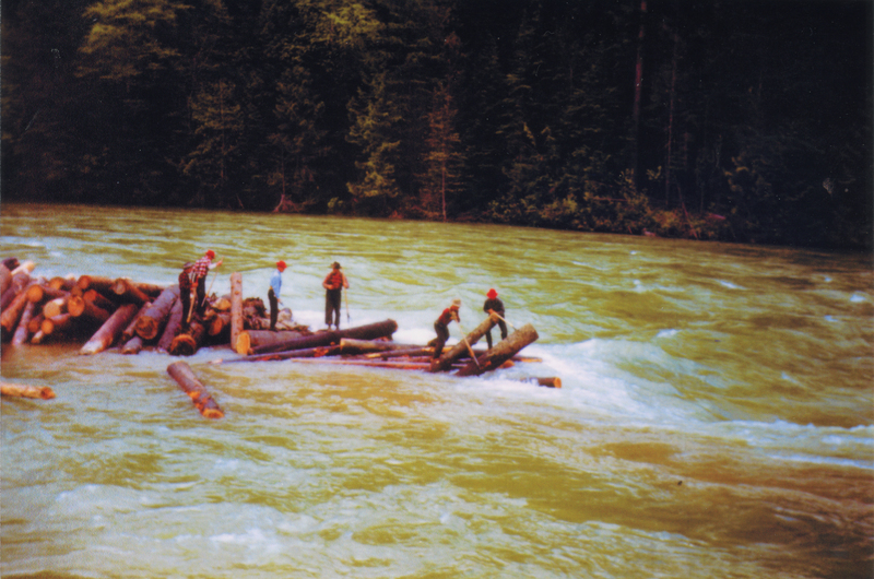 Several individuals wearing helmets and outdoor clothing are standing on a group of logs floating in a river. They are surrounded by flowing water with a forested area in the background.