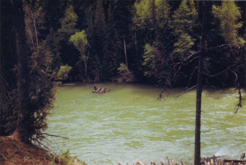 A person standing on a small raft made of logs floating down a river, surrounded by dense trees and forest.