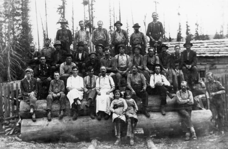 A group of people, mostly men with a few children, sitting and standing on large logs in a forested area. They are dressed in work clothes. In the background, there is a wooden structure and tall trees.
