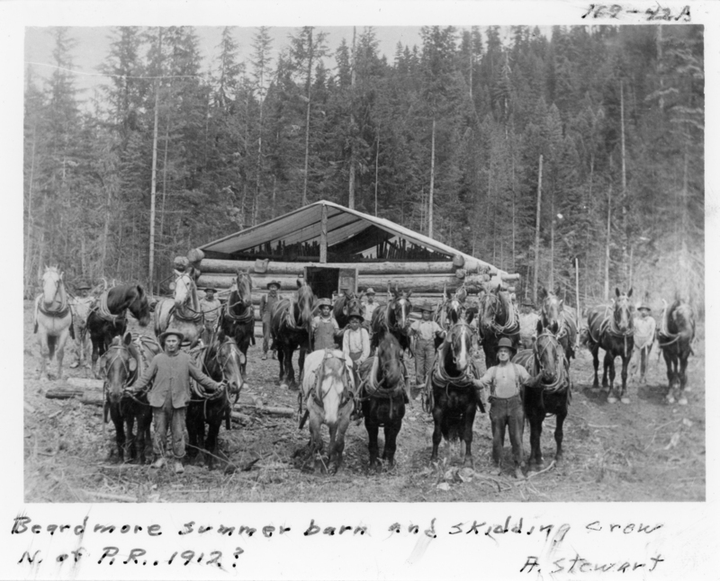 A group of men standing with horses in front of a log barn in a forested area. The barn is rustic, and the men appear to be part of an outdoor crew, possibly for logging or construction. Evergreen trees surround the scene. Handwritten text at the bottom reads: "Beardmore summer barn and skidding crew N. of P.R. 1912? A. Stewart". There is also text near the top right that reads: "129-22A".
