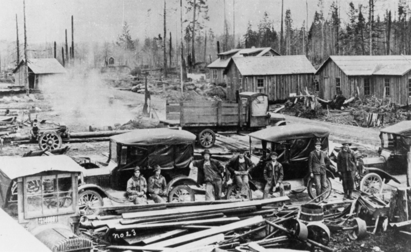 A group of men stand and sit by several early automobiles and in front of a wooden structure. The area is surrounded by cut logs, scattered lumber, and tools, with several wooden buildings and tall trees in the background. Smoke rises from a small fire or machinery in the vicinity. The text "no 23" is visible near the bottom center of the image.