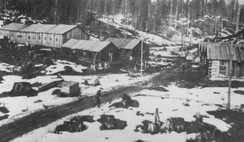 A small settlement in a snowy landscape with several wooden buildings and tree stumps scattered around. A few people are visible walking near the buildings.