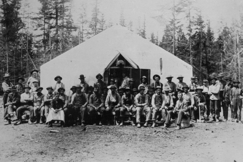 A large group of men posed in front of a white tent in a wooded area. They are dressed in work clothing, with some seated and others standing. The background features tall trees.