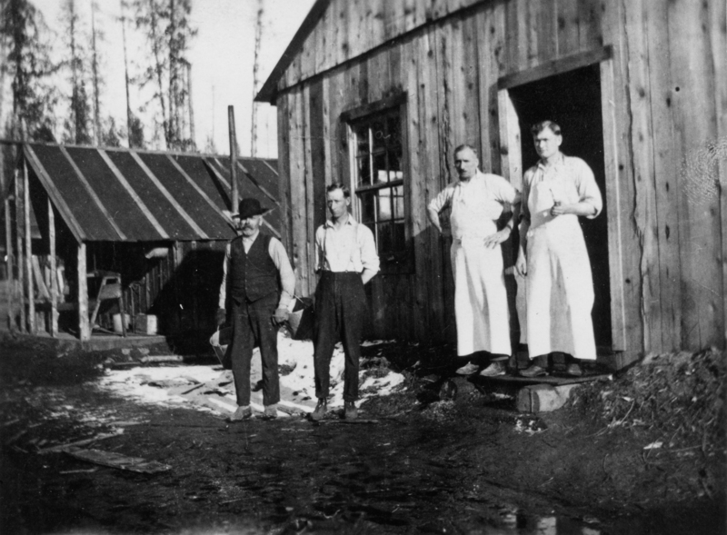 Four men stand outside a wooden building. Two of them wear aprons and are standing near the doorway. Another man in a hat stands holding a small canister. The scene is set in a wooded, rustic area, with another structure in the background. Snow is visible on the ground.