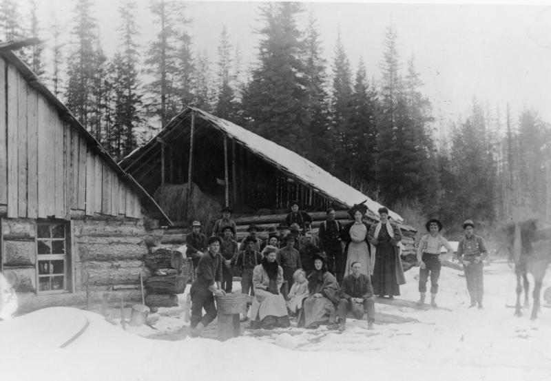 A group of people gathered outside wooden buildings in a snowy landscape with tall trees in the background. Some are seated, while others stand, including a child, wearing a variety of hats and clothing typical of an earlier time period. A horse is visible to the right.