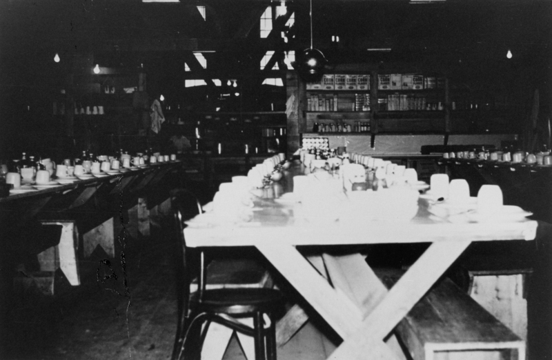 A long table set for a meal with plates, cups, and condiments. The room has shelves stocked with boxes and jars in the background. There are chairs around the table, and hanging light bulbs are visible.