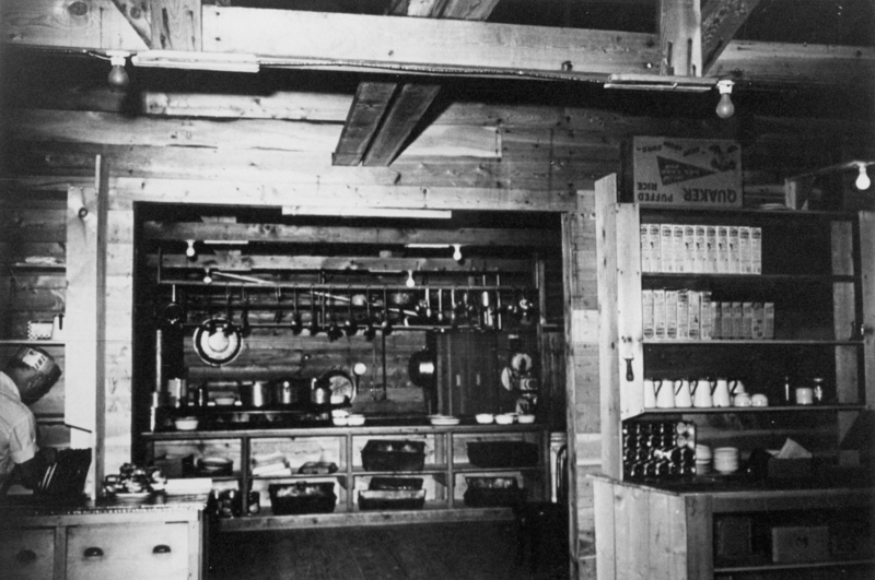 A rustic kitchen with wooden walls and exposed beams. Pots and pans hang above counters filled with bowls and dishes. Shelves hold boxes of cereal and cups. A person stands to the left, working at a counter.
