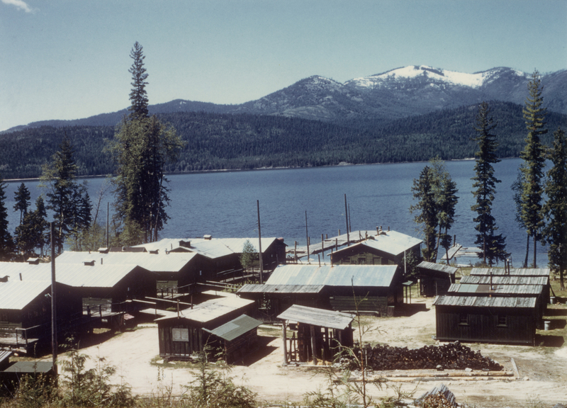 Buildings with metal roofs situated by a lakeside, surrounded by trees, with mountains visible in the background.