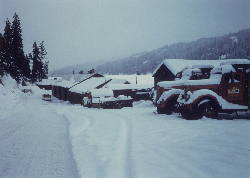 Snow-covered landscape with a road and buildings. Several trucks are parked, also covered in snow. Tall evergreen trees line the hillside on the left, and snow-capped hills are visible in the background.