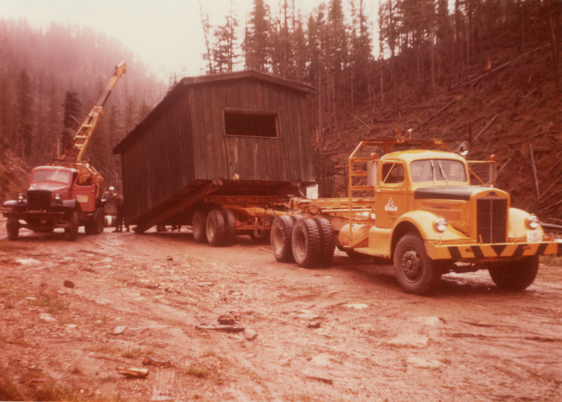 A large, dark wooden structure is being transported on a trailer pulled by a yellow truck. A red vehicle equipped with a crane is positioned nearby. The setting is a wooded, hilly area with several trees on both sides.