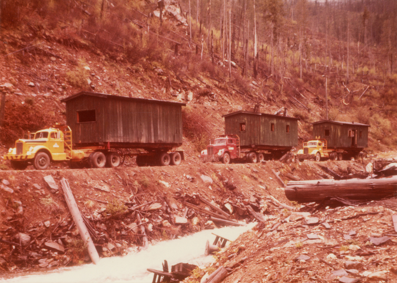 Three trucks are parked in a line on a dirt path. Each truck is carrying a large, dark green cabin-like structure on its trailer. The area around them is a rocky, wooded hillside with sparse trees and debris scattered along a nearby creek.