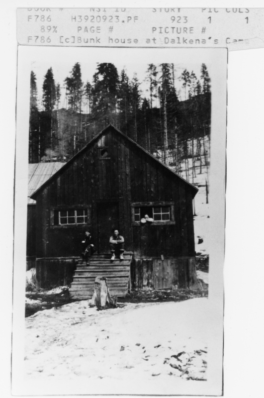 Two people are sitting on the wooden steps of a rustic cabin. A third person is visible through a window. Snow covers the ground, and trees are in the background.