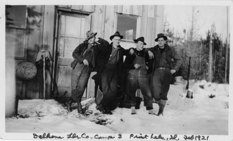 Four men in work clothes and hats standing in front of a wooden building in a snowy landscape, with trees in the background. The men appear to be casually posing, with one resting against another.