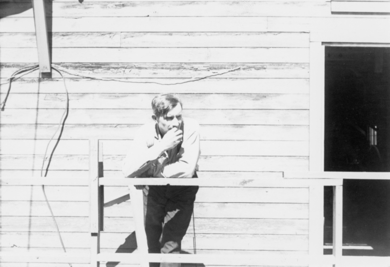 A man leaning on a wooden railing next to a building with horizontal wooden siding. He is holding his hand to his face, looking thoughtful. Part of an open doorway is visible on the right.