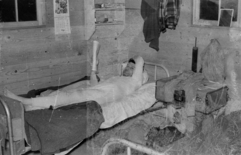 A person lying in a bed with one arm raised, holding a newspaper or magazine. The room has wooden walls with two windows, a calendar on the wall, clothes hanging, and a shelf with small items. There is a suitcase with garments on it beside the bed. Package on shelf reads: "DAM BAR."