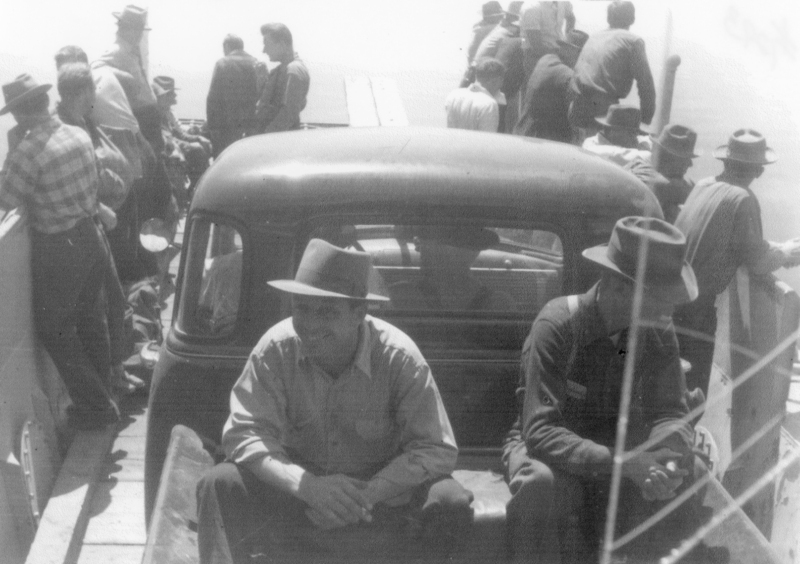 Several men wearing hats are gathered on a wooden platform. Two of them are sitting on the back of a truck, while others stand around, leaning on the sides. The scene appears to be on a boat or ferry, with people facing different directions and some looking out over the water.