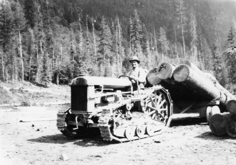 A man wearing a hat operates a tractor pulling large logs in a forested area.