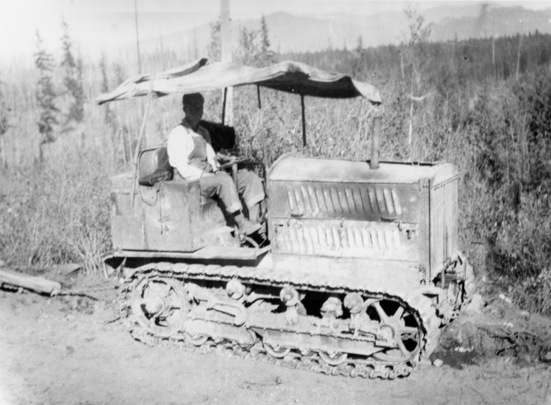 A person sitting on a tractor with tracks, under a makeshift canopy, in a forested area with trees and mountains in the background.