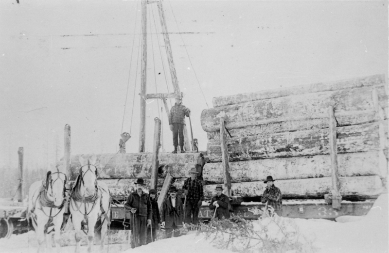 Several men stand in a snowy area next to large logs stacked on a cart. Two horses are harnessed in front of the cart. A man is standing on top of the logs. Near the bottom center of the image on the side of logging bed reads: "NATIONAL" and "33".
